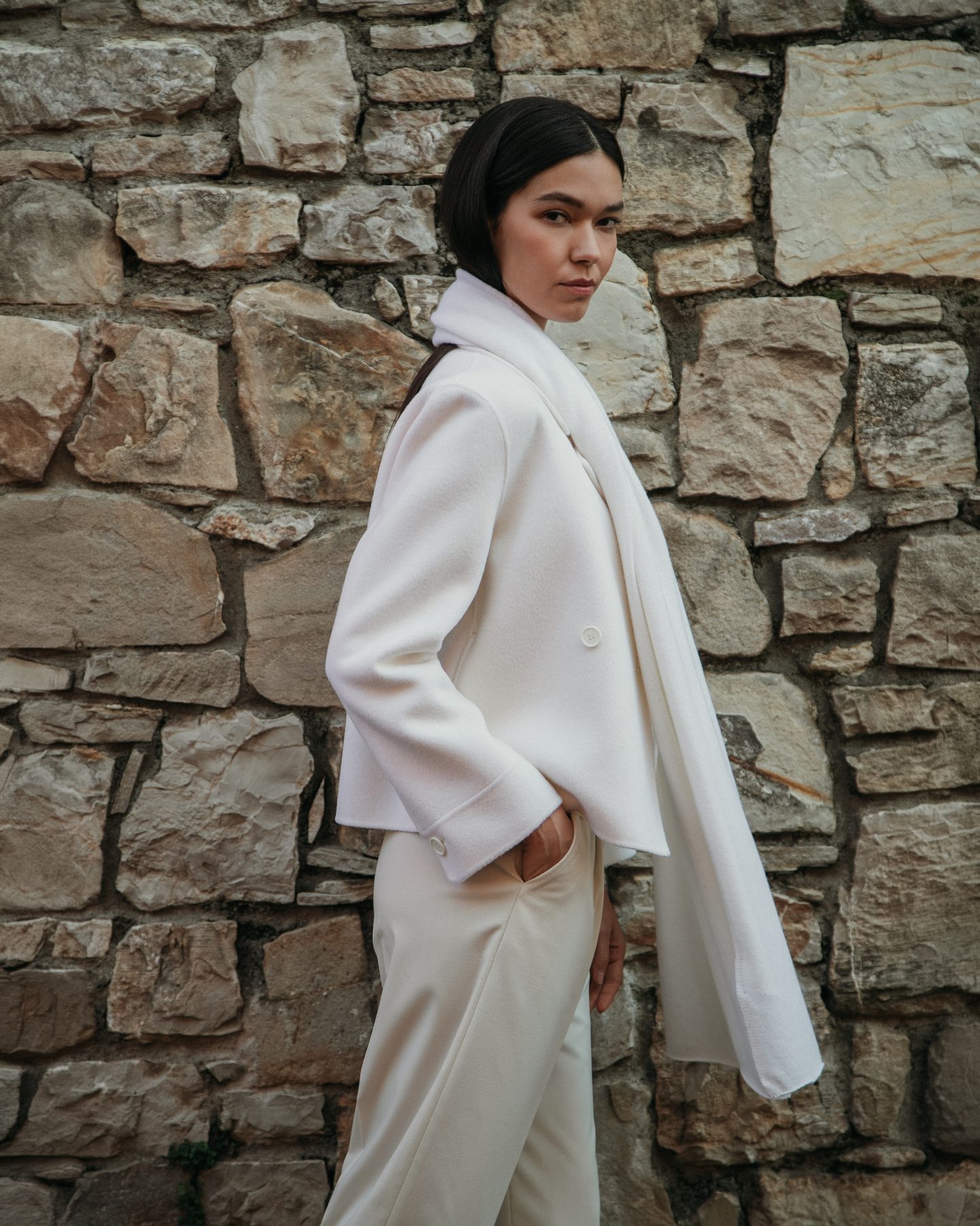 Cappotto e sciarpa in maglia avorio davanti a un muro di pietra